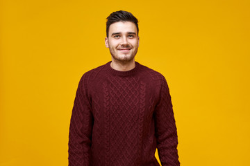 Studio shot of handsome cheerful young man with stylish haircut and dimpled smile posing isolated against blank yellow studio wall background, dressed in cozy maroon sweater, having confident look