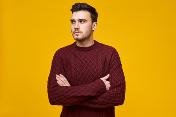 Displeased serious young brunette guy in stylish knitted pullover posing in studio, crossing arms...