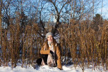 Horizontal full length portrait of pretty fair young woman dressed in warm winter clothes crouching and patting fresh snow during a golden hour afternoon