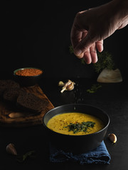 Hot soup with red lentils, spices, herbs, vegetables and cream. Soup on a blue napkin, next to brown bread, garlic, a bowl with lentils. Female hand adds spices to the soup. Close-up.