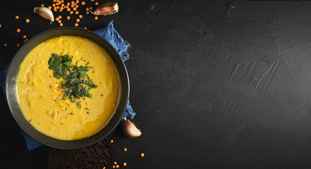 Red lentil soup, spices, herbs, vegetables, lemon and cream. Soup on a blue napkin, next to garlic cloves. Lentil seeds are scattered on a black background. Top view. Copy space for text.