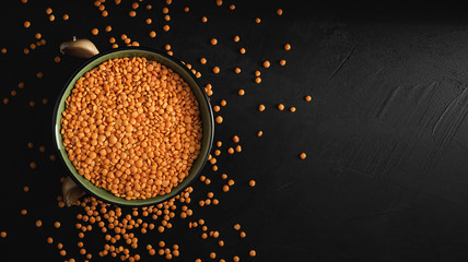 Bowl with seeds of red lentils on a black stone background. Lentil seeds and cloves of garlic are scattered next to the bowl. Flat lay. Copy space for text.