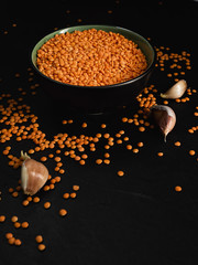 Bowl with seeds of red lentils on a black stone background. Lentil seeds and cloves of garlic are scattered next to the bowl.