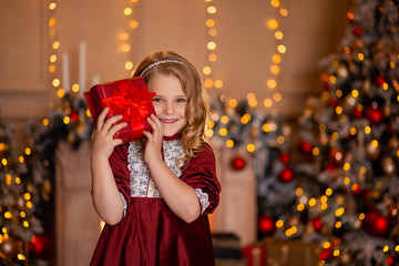 A cute girl with blond hair in a red elegant dress laughs cheerfully and holds a gift box in her...
