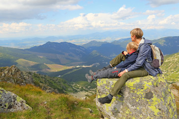 Naklejka na ściany i meble Mother and daughter sitting on large stone and watching mountain view