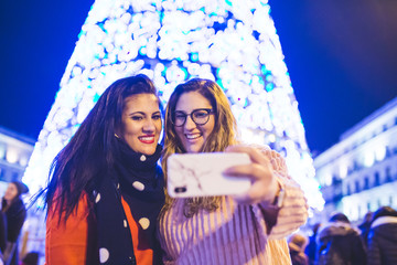 Two girls are taking a picture next to the Christmas lights. They are enjoying together in the city...