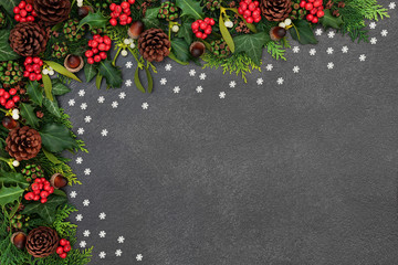 Natural Winter Christmas & New Year background border with  holly, ivy, acorns, pine cones, cedar leaves & mistletoe with loose snowflake decorations on grunge grey background with copy space.