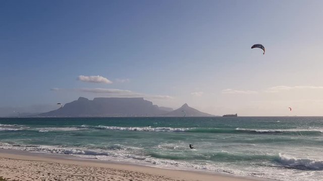Panoramic view of Table Mountain and Cape Town in windy conditions. Kitesurfers and kiteboarders busy kitesurfing on choppy seas. Big ship on horizon. Waves breaking on Bloubergstrand beach.