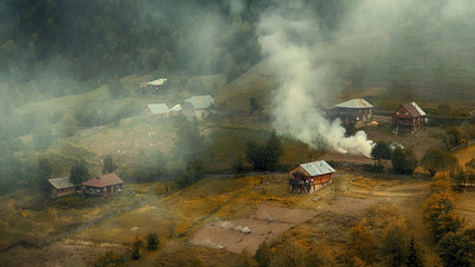 Fototapeta premium Villagers burn foliage and grass in their gardens in the fall, smoke and fog rises above the village, hills and mountains.