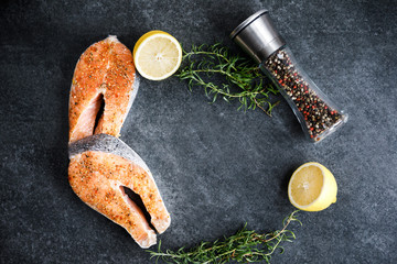 Fresh salmon steaks lie on a dark background, alongside lemon and rosemary and peppercorns with different types of peppers. Top view
