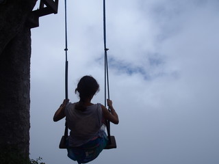 A young woman sitting on a large wooden swing at the treehouse under a cloudy sky, La casa del Arbol (La Casa del &Aacute;rbol), Banos (Ba&ntilde;os), Ecuador
