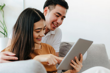 young asian love couple talking and using mobile tablet, watching movie online on sofa at home office, internet technology, social media, online booking, entertainment, shopping online concept