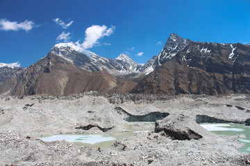 A beautiful lake of melted ice in the middle of Ngozumpa glacier strewn with stones. Chola mountain rises above it. Clear and blue sky with some clouds. Route to Everest base camp through Gokyo Lakes.