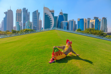 Travel in Qatar, Middle East. Happy woman in sunhat sitting on a green lawn in a park with modern...