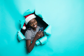 Obraz premium Young african woman wear in santa hat use phone in hands standing in green torn paper hole background