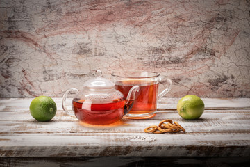 Still life on a light textured background glass teapot mug with poured tea limes on a table of...