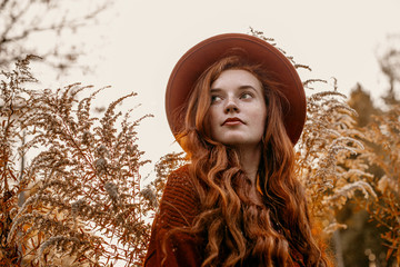 Outdoor close up portrait of young attractive redhead girl with natural freckled skin, long curly hair, wearing stylish orange hat, posing in beautiful autumn nature. Copy, empty space for text