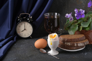 Breakfast. Boiled egg on a white stand, a glass of tea, rye bread, an alarm clock and a pot with a violet flower on the table.