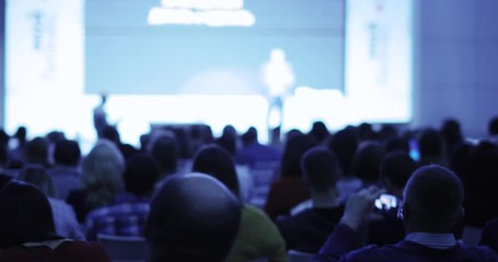 Unrecognizable businessman on stage talking and showing presentation on screen to large audience sitting back to camera, blur background. Training, conference, public speech concept.