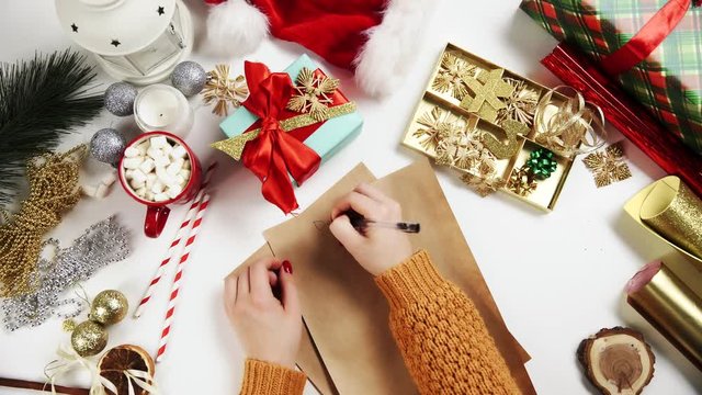 Female hand writing a gift list on wooden table with gifts, wrapping paper, red Christmas decorations.