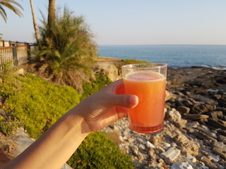 Orange juice in woman hands on the beach with sea and palms background. Girl is holding a red cocktail with a slice of orange.