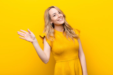 young pretty blonde woman smiling happily and cheerfully, waving hand, welcoming and greeting you, or saying goodbye against flat color wall