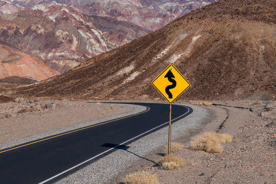 Naklejki A crooked road sign along a highway curving through a barren and rugged desert landscape