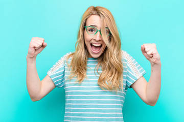 young pretty blonde woman feeling happy, surprised and proud, shouting and celebrating success with...