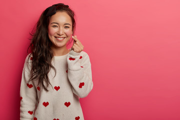 Romantic Chinese woman shows korean heart sign with two fingers crossed, smiles joyfully and...