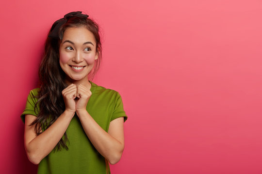 Attractive Asian teenage girl smiles tenderly, keeps hands together, eager to get surprise, grins happily, looks aside, has dark pony tail, wears green t shirt, isolated over pink background