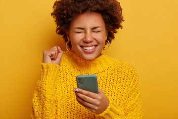 Close up shot of overjoyed curly woman keeps fist clenched, happy to get money reward, gets notification on cellphone, wears yellow knitted sweater, stands indoor, feels amused and entertained