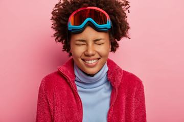 Winter holidays and vacation concept. Close up shot of joyful Afro American woman wears ski goggles, enjoys leisure time, dressed in turtleneck and red jumper, isolated on pink wall. Happy skier