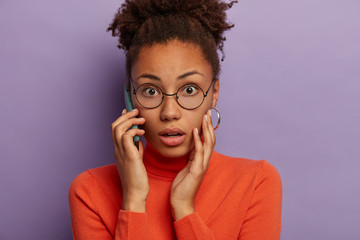 Headshot of stunned emotional dark skinned woman gasps from fear, hears unpleasant news, has phone conversation, wears optical glasses and turtleneck, models over purple background. Omg concept