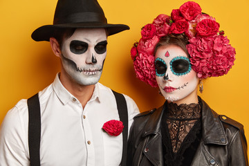 Calm young woman and man wears skull makeup, female in beautiful floral wreath, dressed in...