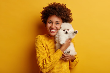 Photo of pleasant looking Afro woman holds cute small snow white spitz near face, prepare for dog party, cares very well about pet, ready for strolling, express faithful feelings. Tight embrace