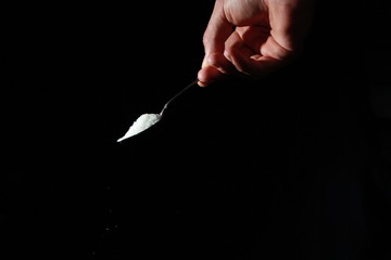A male hand holds a small spoon from which white matter is poured out in total darkness under studio lighting