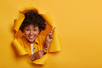 Positive Afro American lady shows something amazing, points up and sideways with index fingers, advertises copy space, has toothy smile, isolated over yellow background, poses in paper hole.