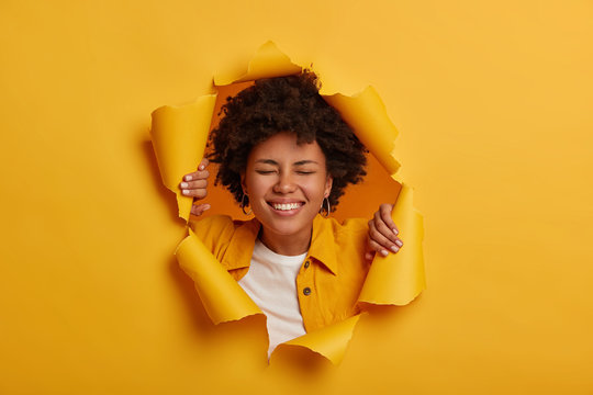 Lucky overjoyed African American woman smiles broadly, has carefree mood, eyes shut, dressed in fashionable clothes poses in yellow paper background with ripped hole being full of energy chills indoor