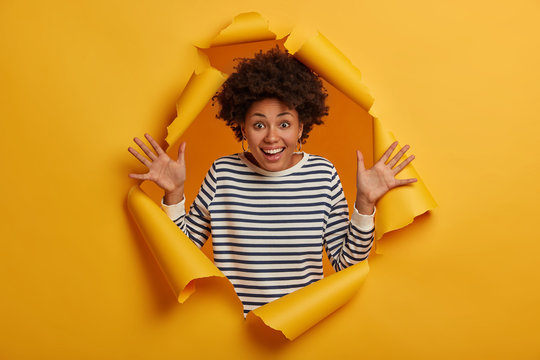 Photo of joyful curly woman raises palms, rejoices final meeting with close friend, dressed in striped casual jumper, gestures from happiness, poses in torn ripped paper background, being amused