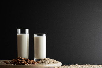 Two glasses of oat milk and almond milk with oat flakes and almond nuts on wooden disk. Copy space, black background.