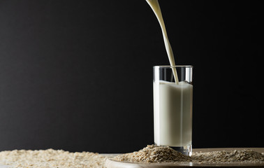 Pouring oat milk into glass on black background and oat flakes on table. Oat milk lactose free milk...