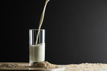 Pouring oat milk into glass on black background and oat flakes on table. Oat milk lactose free milk...