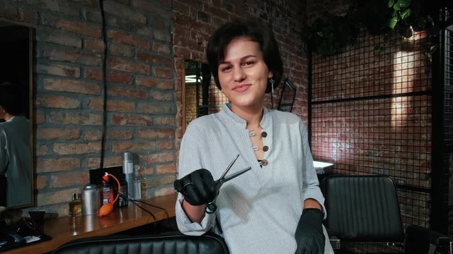 Portrait of smiling beautiful woman hairdresser looking at camera while holding professional scissors on salon background. Beauty and people concept