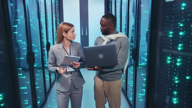 Woman IT administrator with chief african server engineer coworking in cyber security walking through server room discussing issues using a laptop. Professional team.