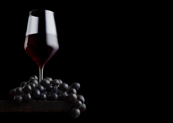Elegant glass of red wine with dark grapes on wooden board on black background. Space for text