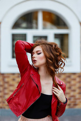 Young curled girl weared in red leather jacket on background of white arch window. Fashion style...