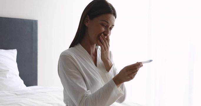 Excited young lady holding positive pregnancy test sit on bed