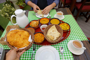 Traditional Bahrain breakfast delicious set served by woman hand on table with man hands wrapping curry in roti on plate with chai tea mixed with milk in restaurant background.