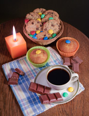 una pequeña taza de café en un plato con bombones confitados sobre un mantel a cuadros blanco y líneas azules amarillas y rojas en una mesa de madera marrón con dulces y bombones, una vela cuadrada na