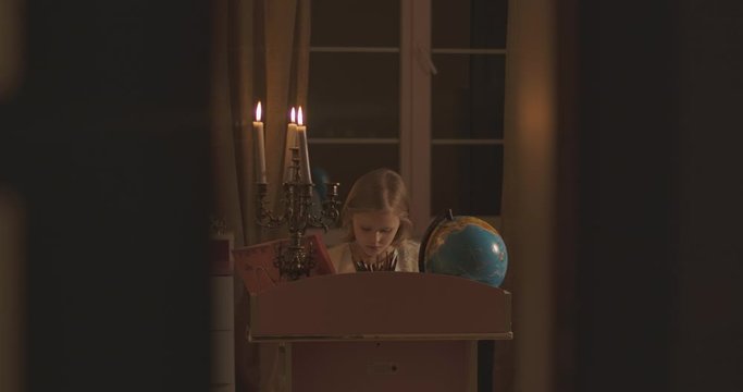 Little Caucasian schoolgirl sitting at the desk and doing homework in candlelight. Blond child hearing something frightening and looking around scaredly. Loneliness, fear, scare.